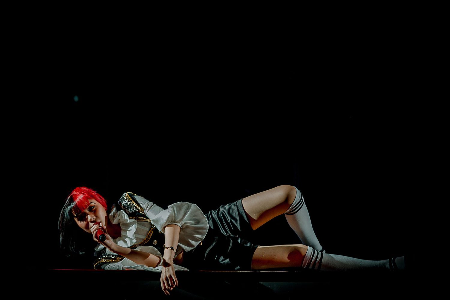 Performer in red hair and dark outfit lying dramatically on stage under theatrical lighting.