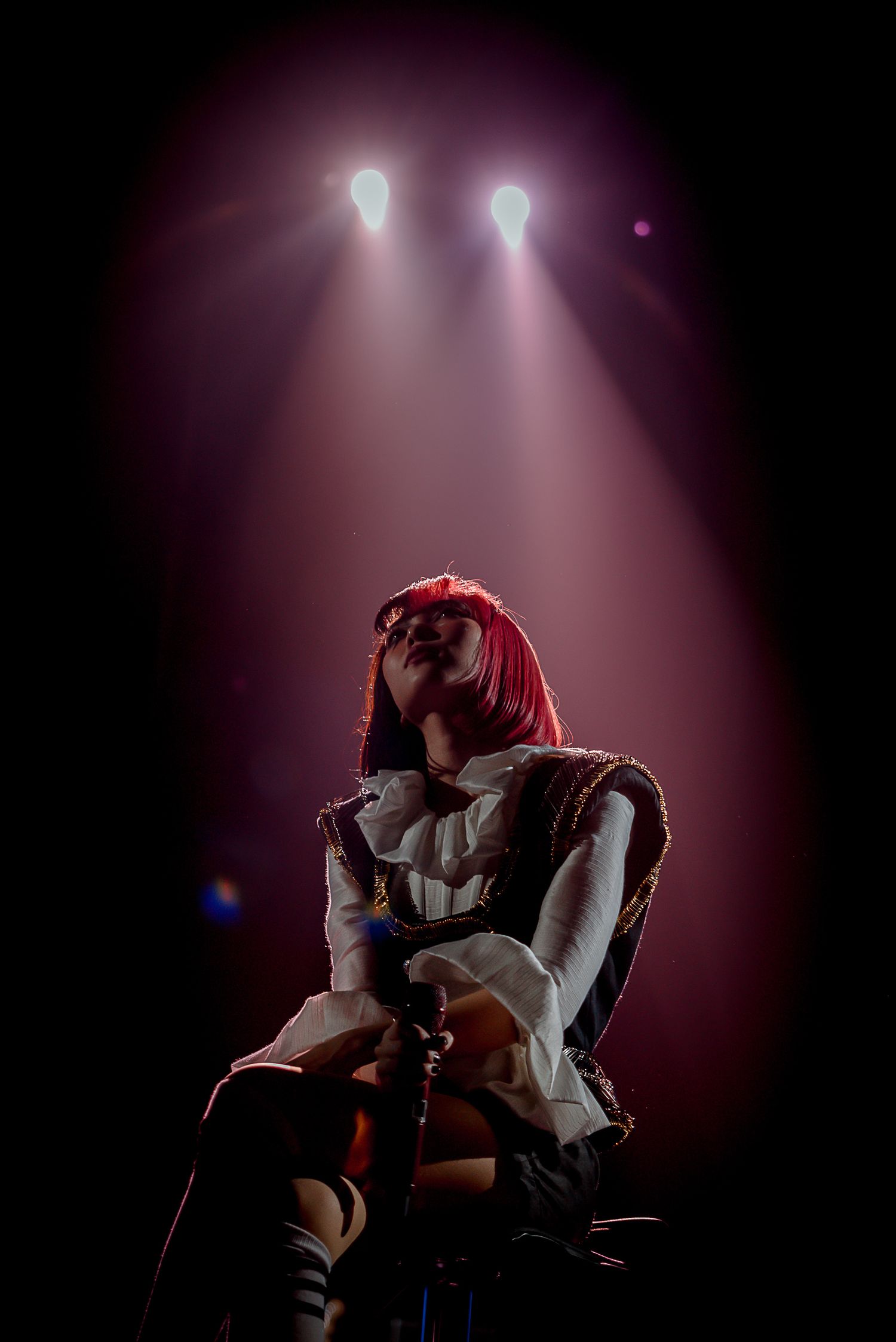 Dramatic stage performance with red-haired figure under dual spotlights creating atmospheric effect.