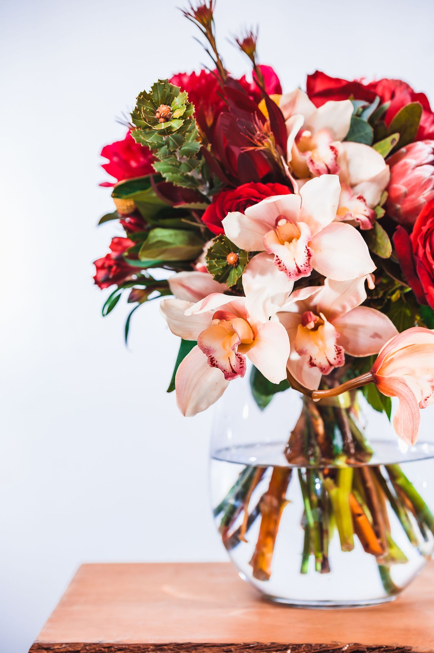 Fresh bouquet featuring pink cymbidium orchids and red roses in a round glass vase on wooden surface.