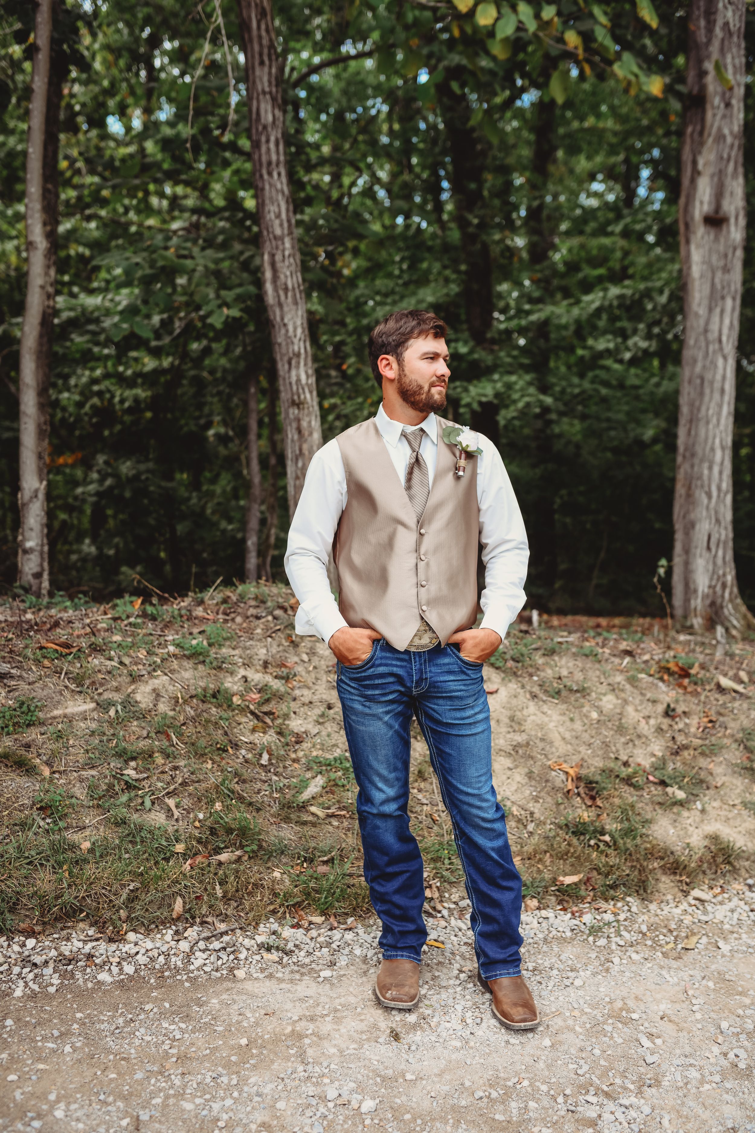 Groomsman in vest and jeans poses casually against wooded background.