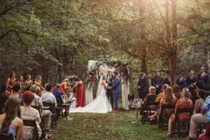 Outdoor wedding ceremony in wooded area with guests seated on rustic benches.