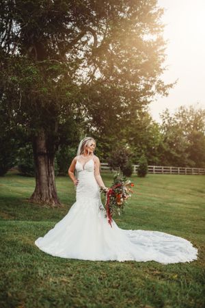 Bride in flowing white dress poses in a sunlit garden setting with red bouquet.