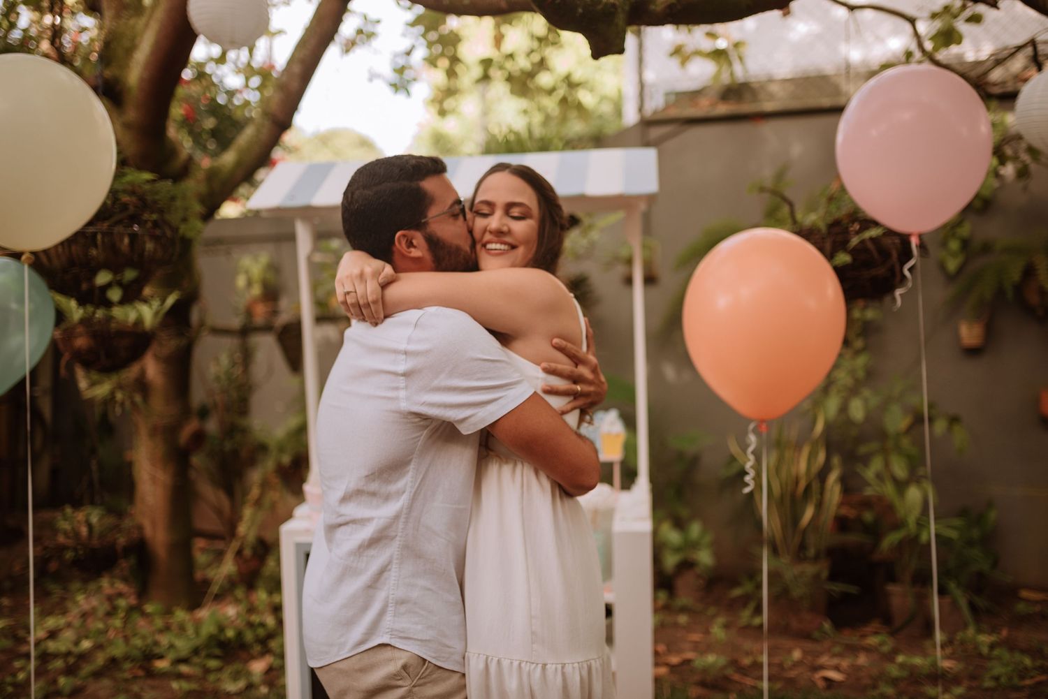 A couple embraces in an outdoor setting with pink and white balloons in the background.