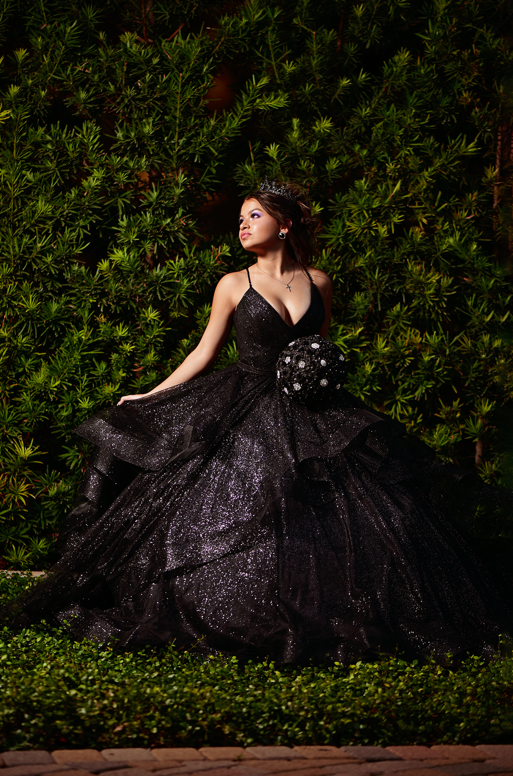 A glamorous black ball gown sparkles against lush greenery in an elegant outdoor portrait setting.