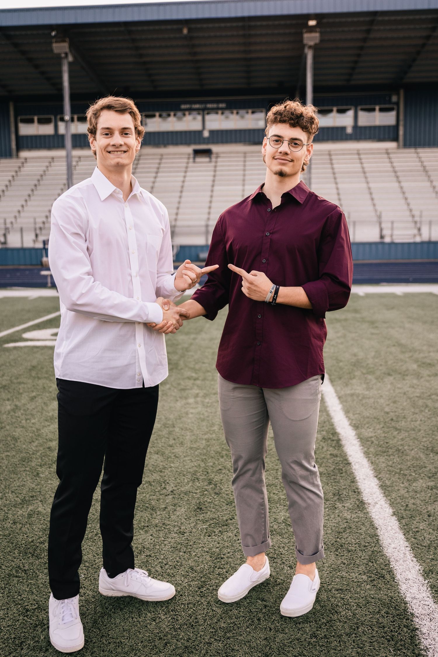 Two young men in casual attire shaking hands on sports field with stadium seating behind them.