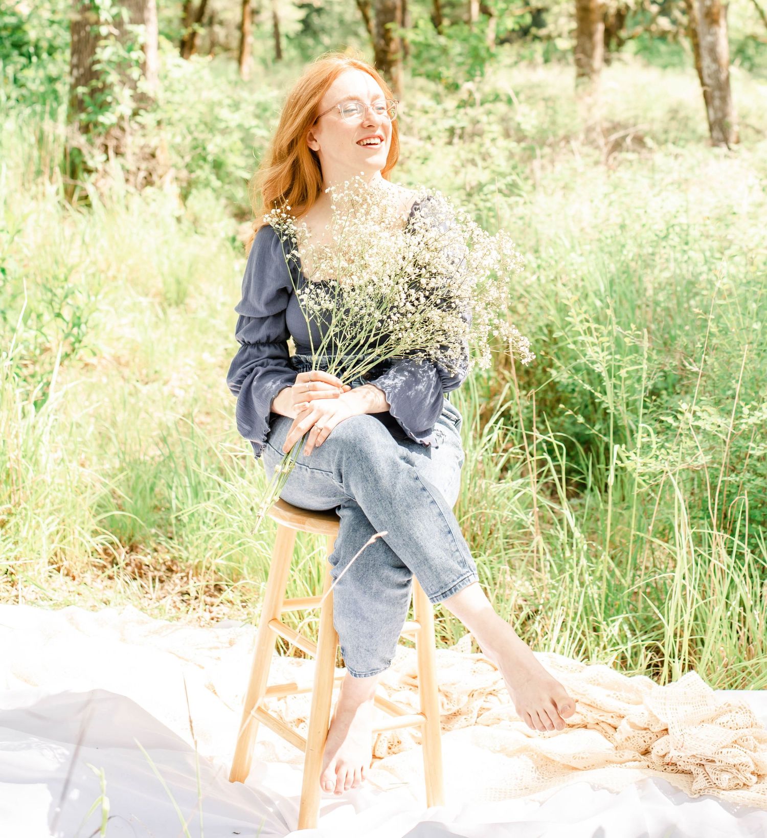 Person in blue dress sits among tall grass with white fabric and wooden stool outdoors.