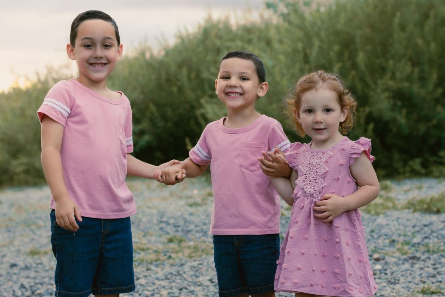 3 kids with matching outfits. older brother, middle brother, and younger sister. Holding hands. smiling at camera. river