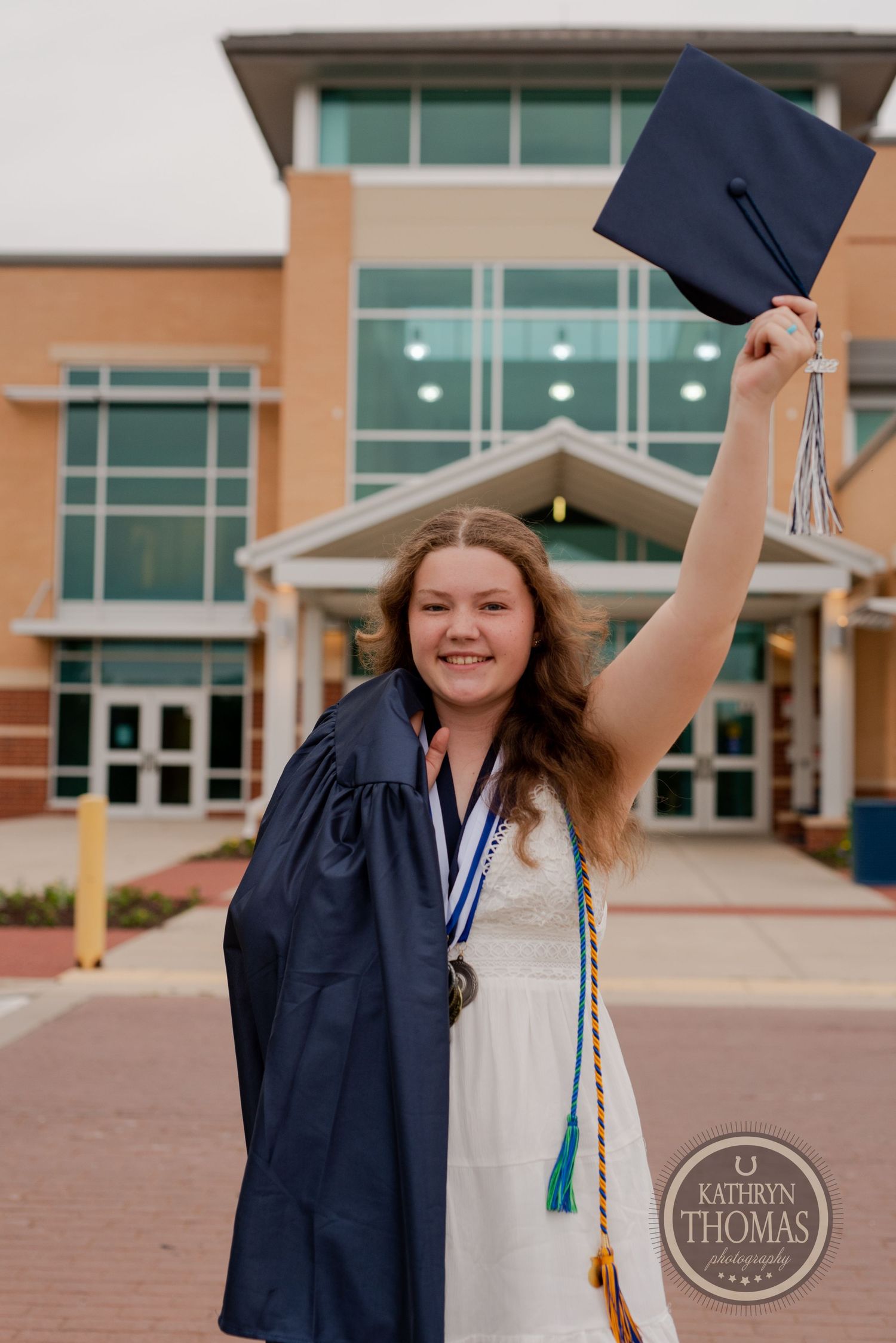 Graduate celebrates by raising cap in the air while wearing white dress outside school building.
