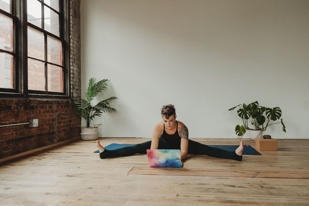 Yoga practitioner in splits position using laptop on wooden floor in minimal studio space.