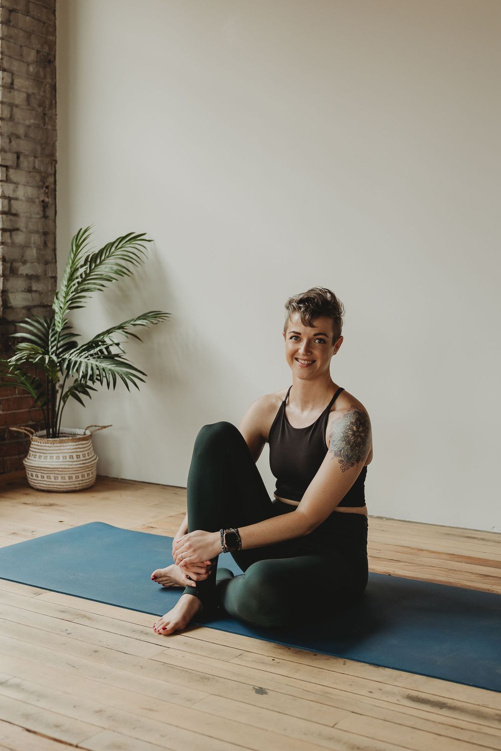A person in black workout clothes smiles while sitting on a blue yoga mat in a studio setting.