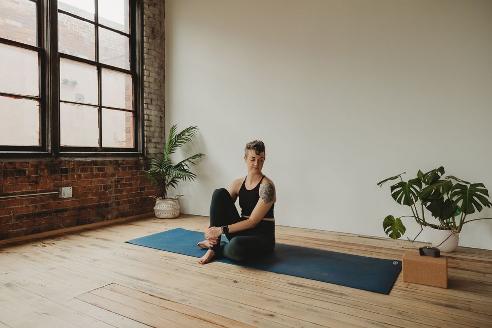 A person sits in a meditative pose on a yoga mat in a bright studio space with brick walls and houseplants.