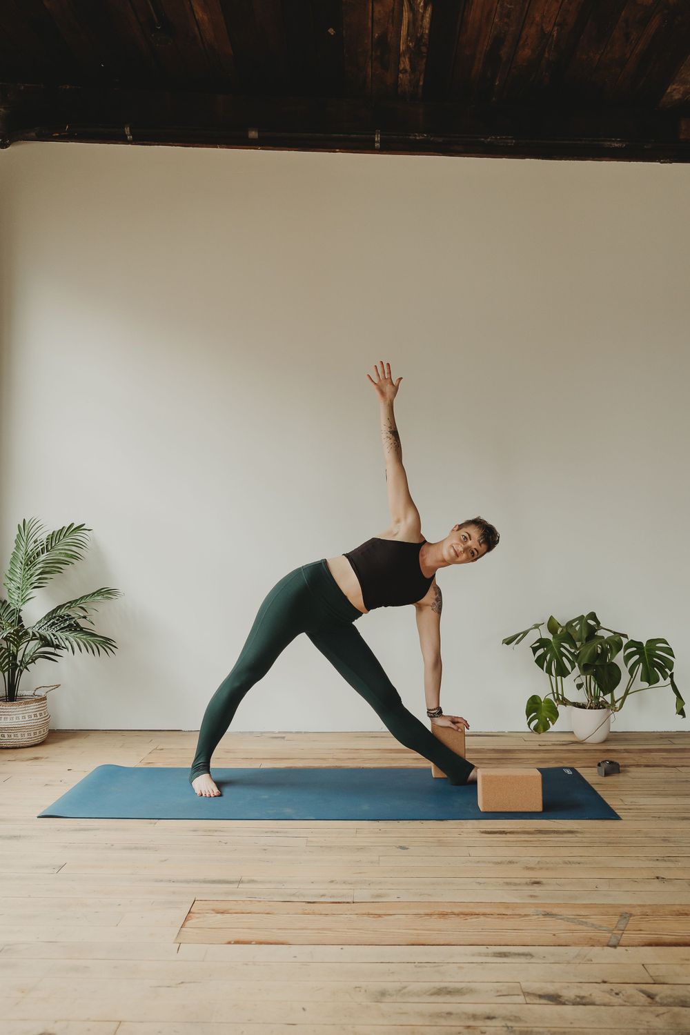 Triangle pose being demonstrated using a wooden block for support on blue yoga mat with plants in background.