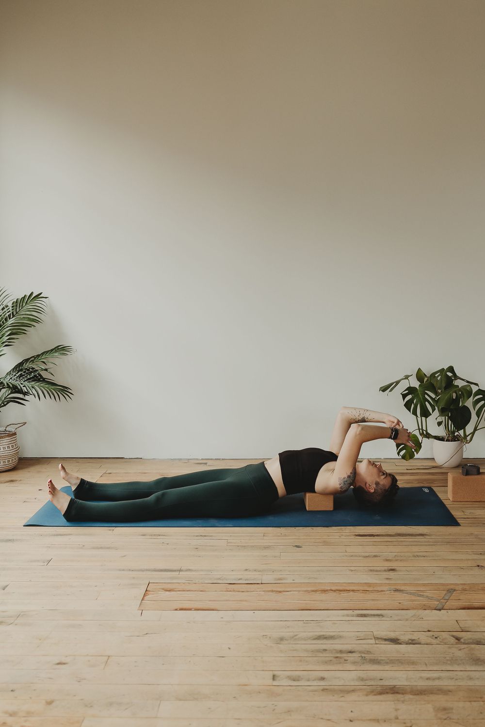 Restorative yoga pose demonstrated on blue mat with wooden block under head in serene studio space.