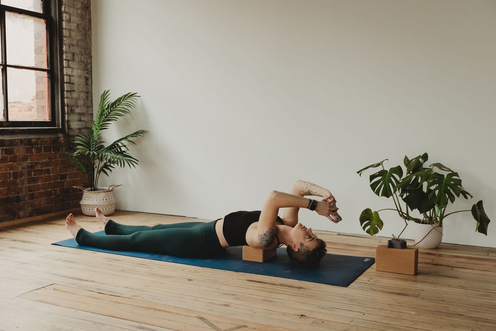 Supine yoga pose shown on blue mat with wooden block support in studio with brick walls and plants.