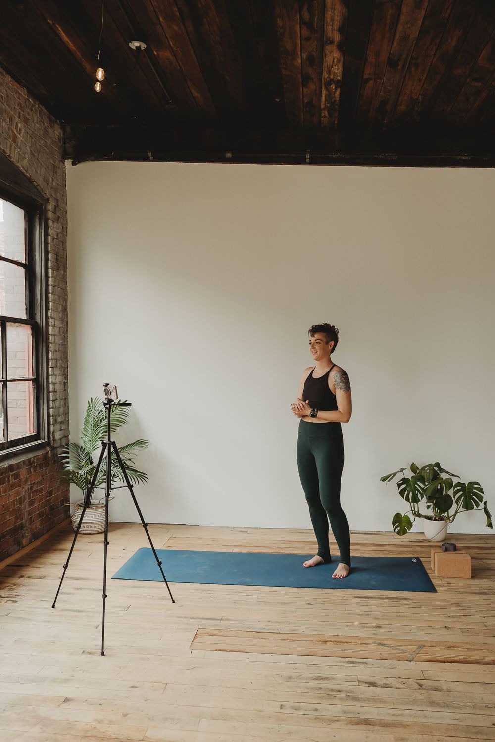 Fitness content creator standing on yoga mat next to camera tripod in industrial-style studio space.