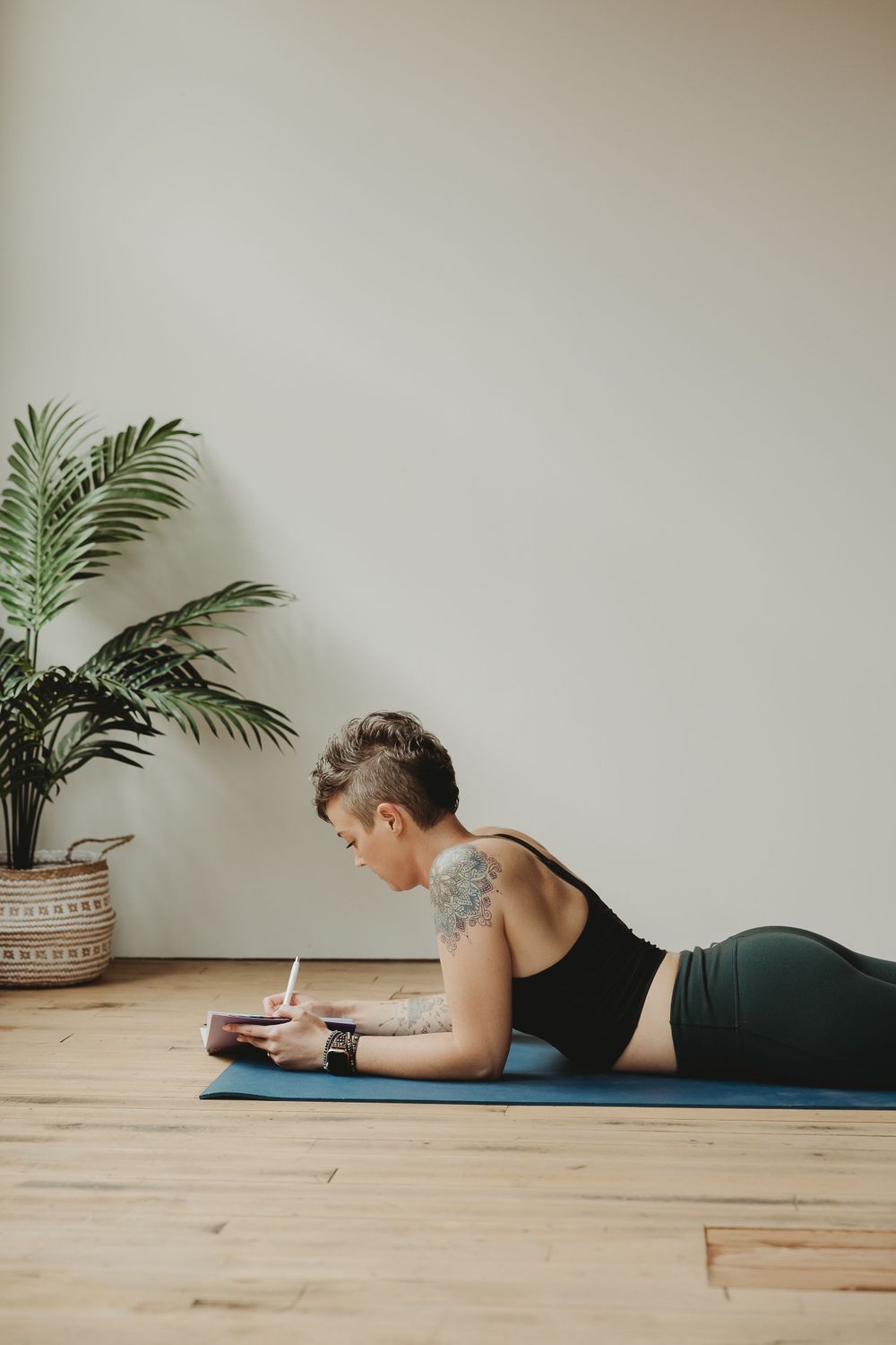 Person writing in journal while lying on exercise mat with decorative palm plant in background.