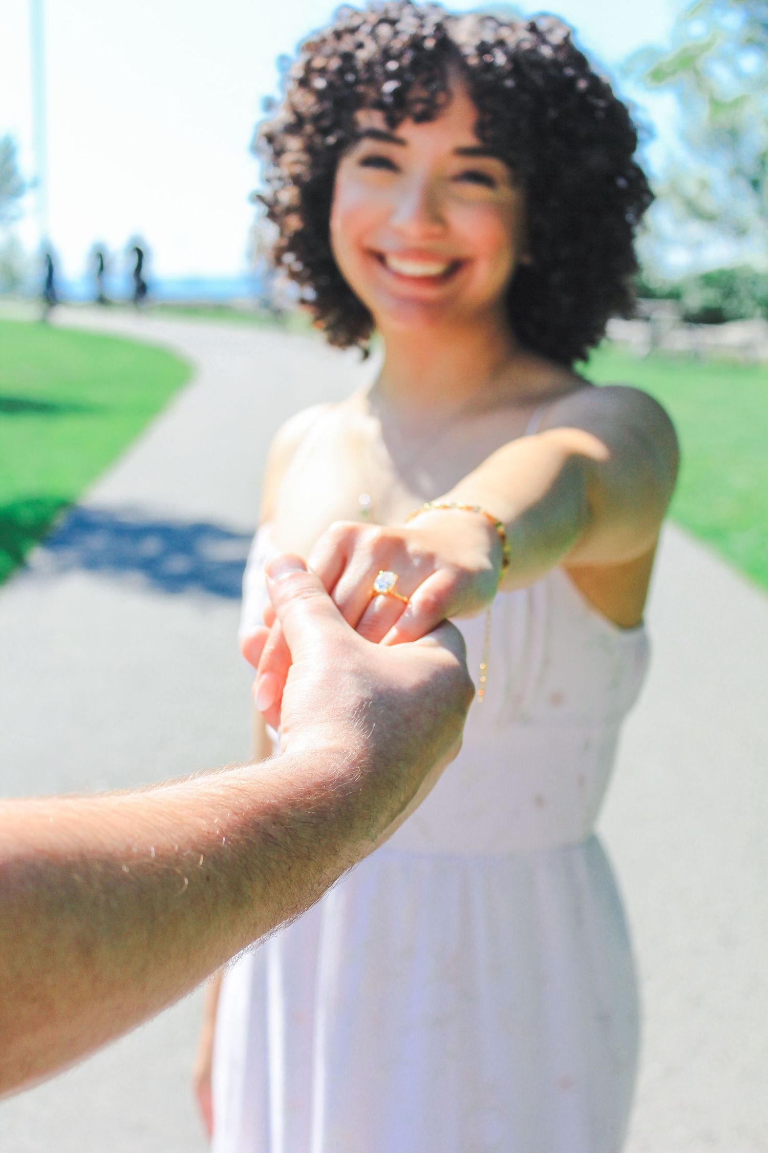 A close-up engagement photo showing a ring as someone holds their partner's hand on a sunny day.