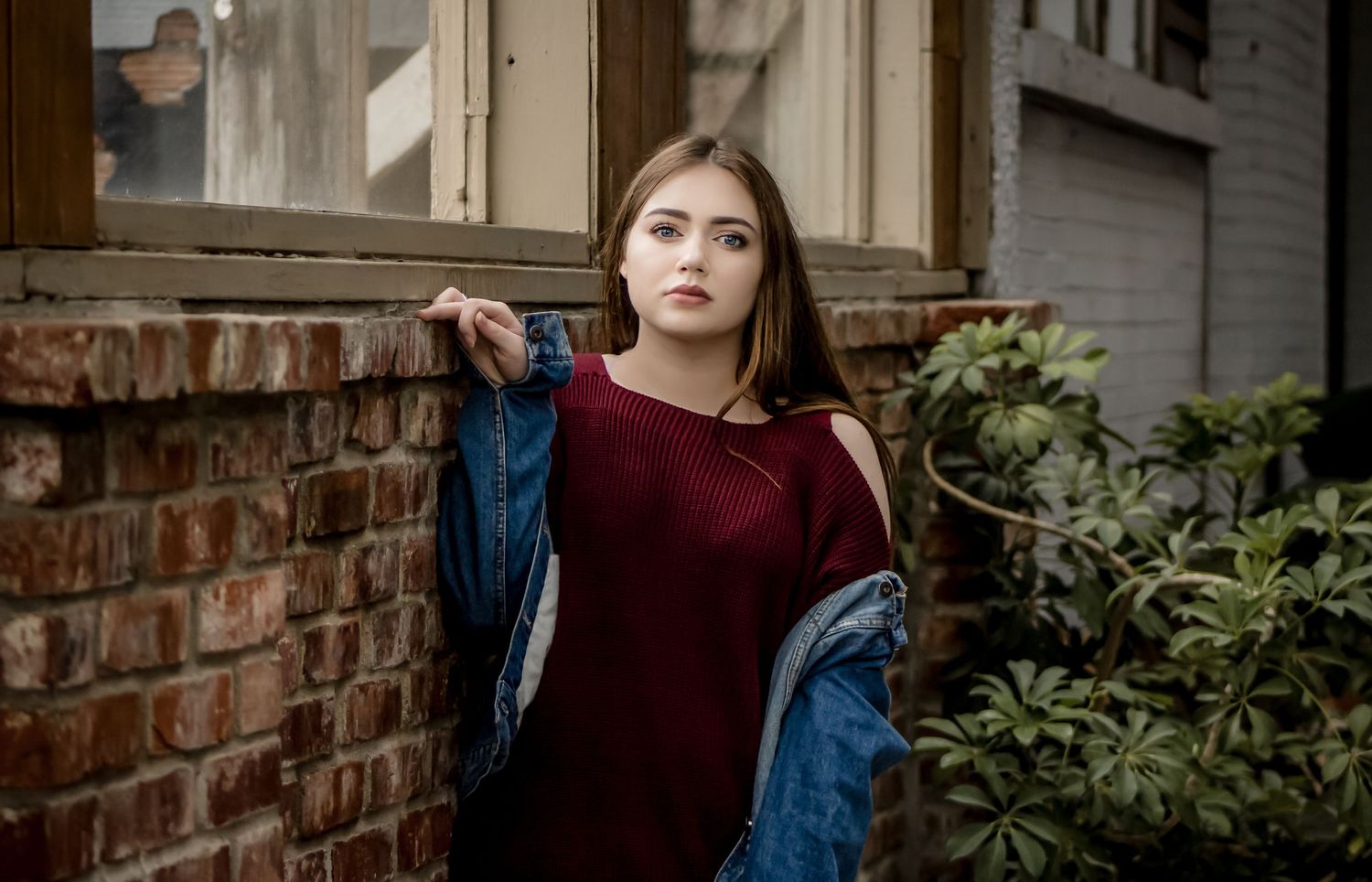 Young woman in red dress and denim jacket posing against a brick wall outdoors.