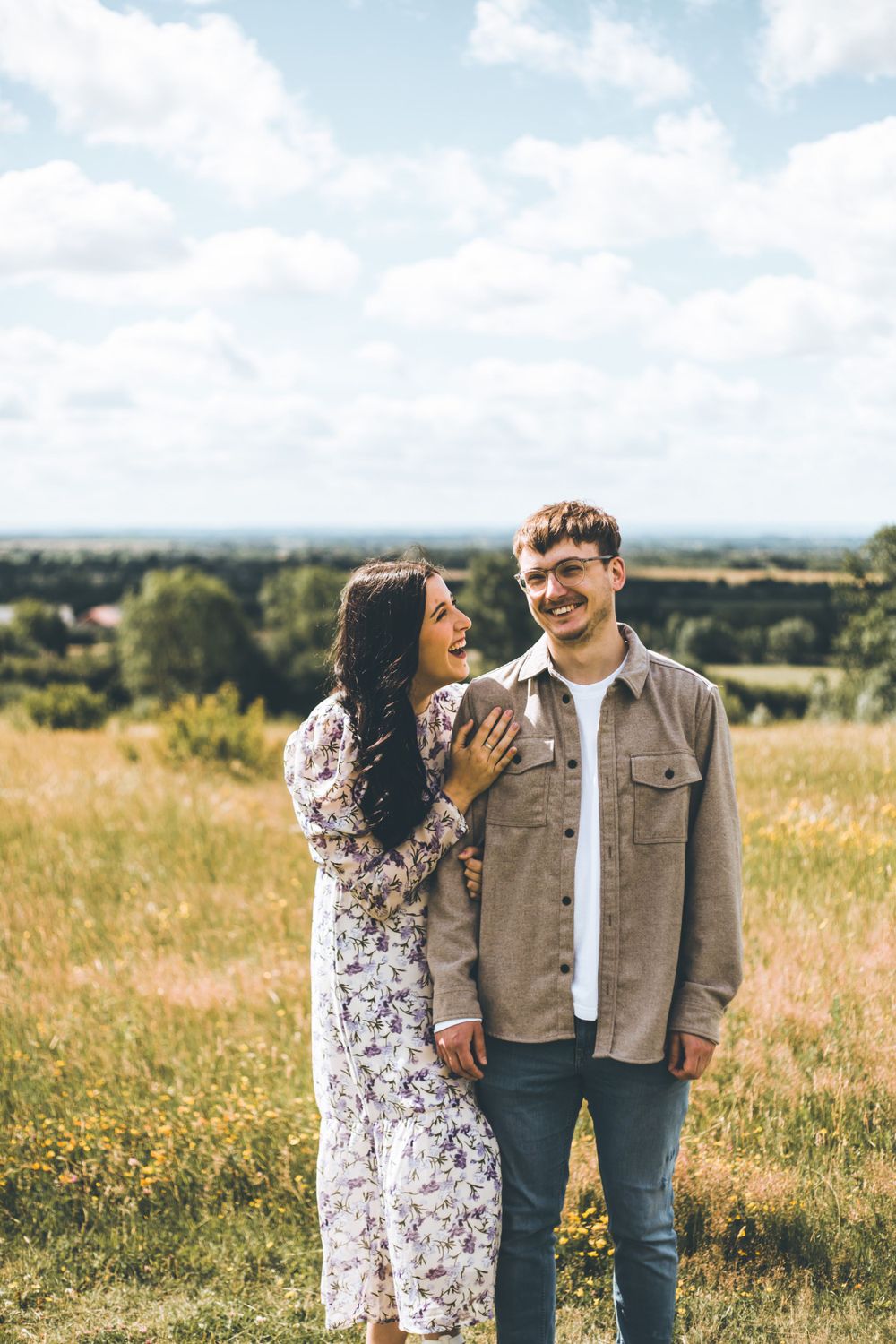 A couple sharing a cheerful moment in a sunny meadow with hills in background.