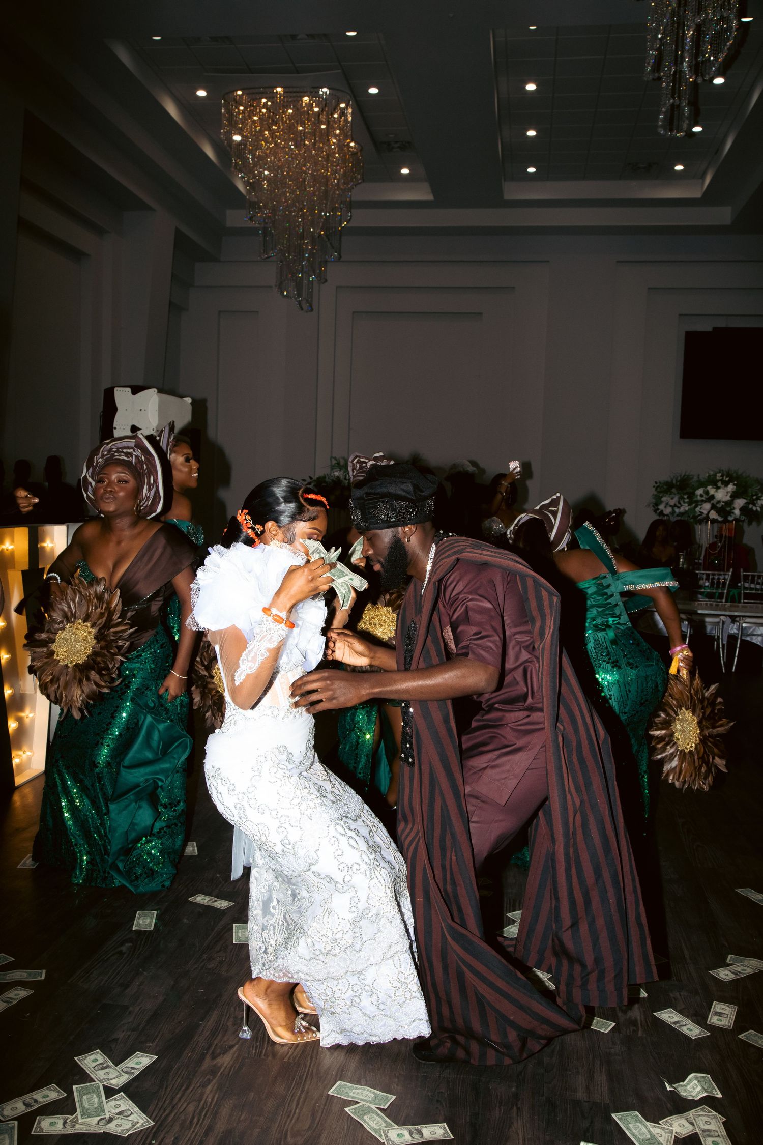 Wedding celebration with people dancing under a crystal chandelier with money scattered on the floor.
