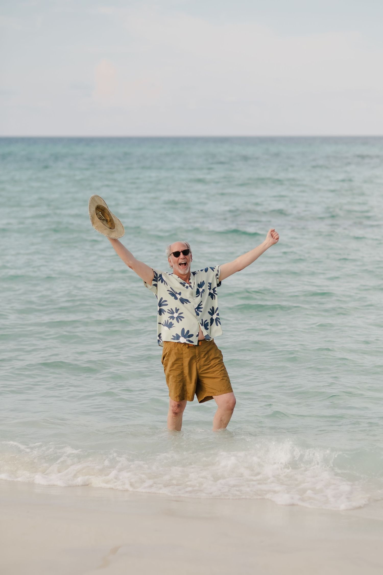 Person in floral shirt and shorts jumping joyfully on a sunny beach.