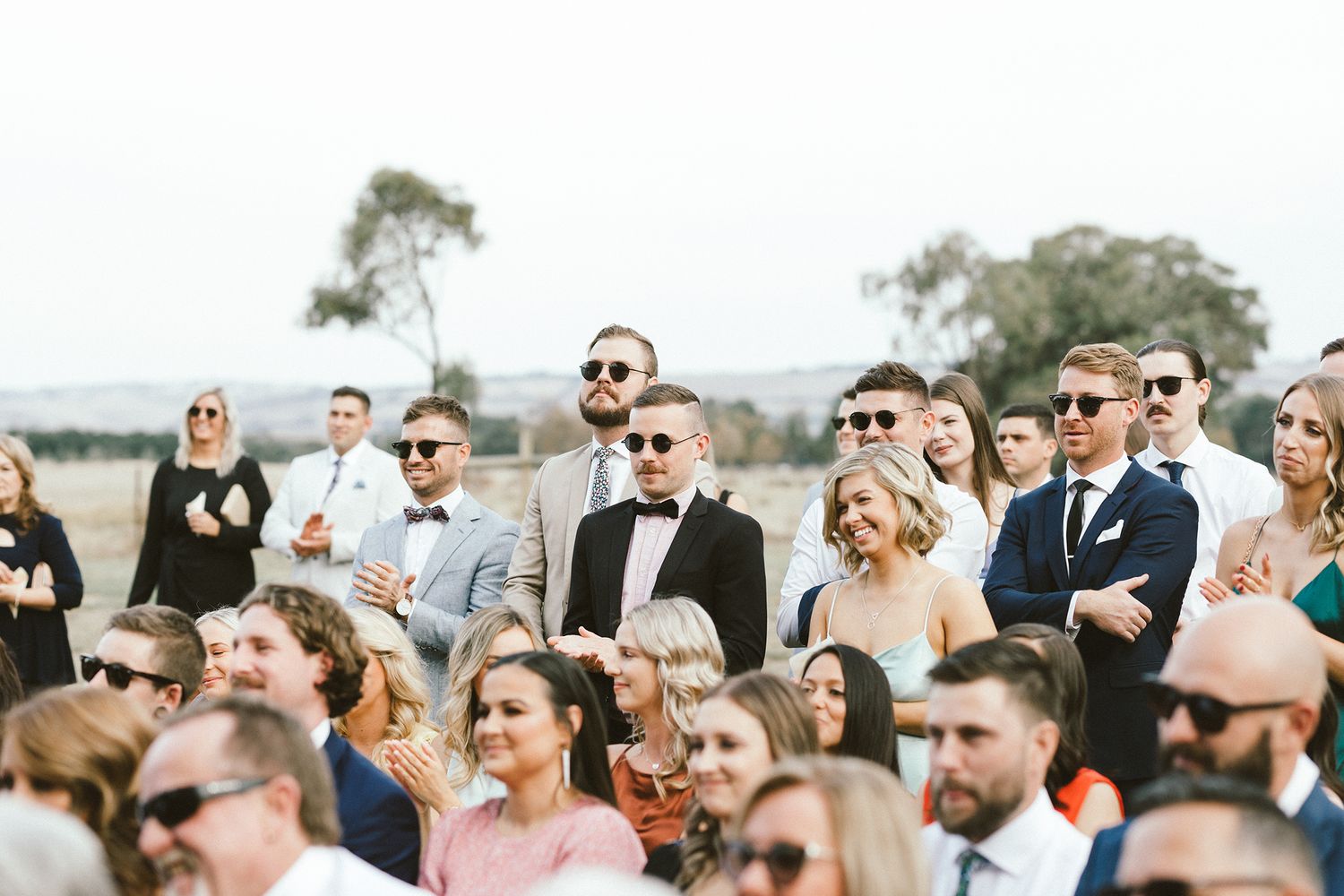 Wedding guests wearing sunglasses watch the ceremony from white chairs outdoors.