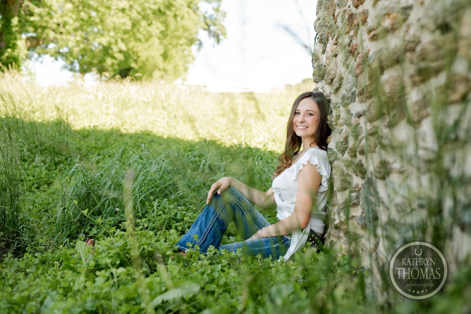 A woman in casual clothing sits by a stone wall surrounded by green grass.