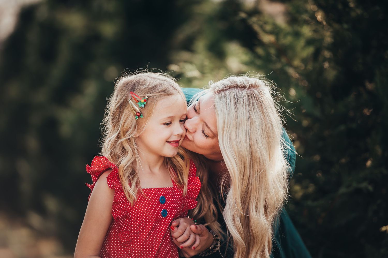 blonde mom leans down to kiss the cheek of daughter with christmas clips in her blonde hair