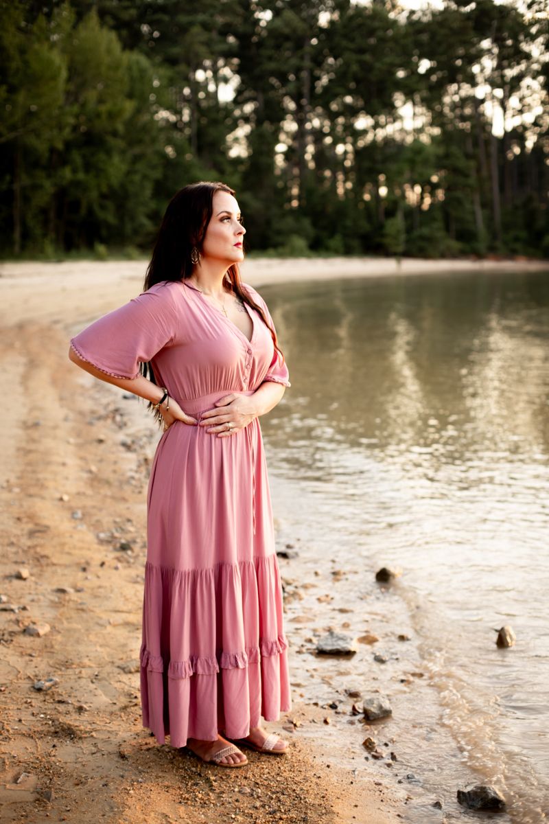 Woman in dusty rose dress on the shore of the lake looking out over the water.