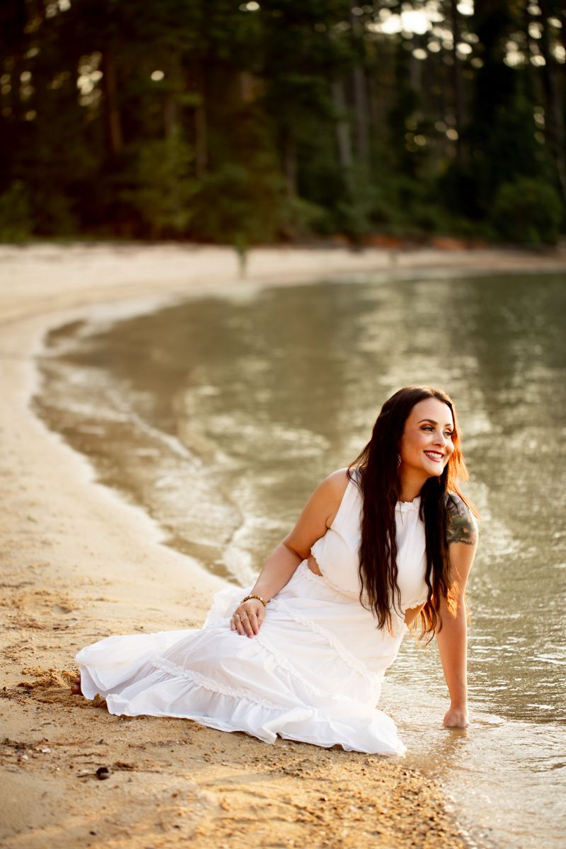 Woman in white dress sitting on the shore of Clarks Hill Lake, SC, looking out over the water smiling.