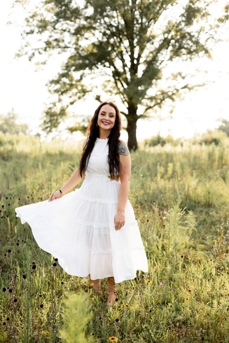 Woman in white dress in a grassy field with sun coming through oak tree behind her.