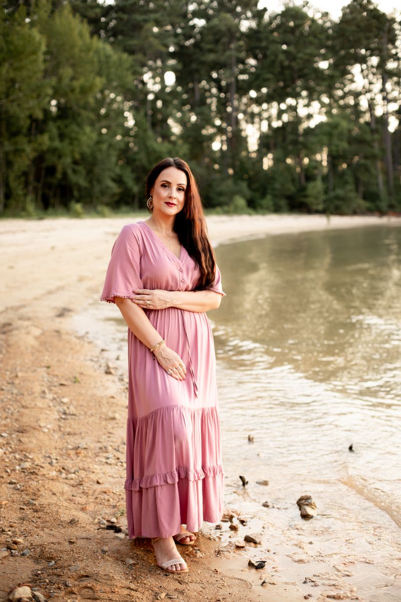 Woman in dusty pink dress standing on the shore of Clarks Hill Lake, SC looking at the camera.