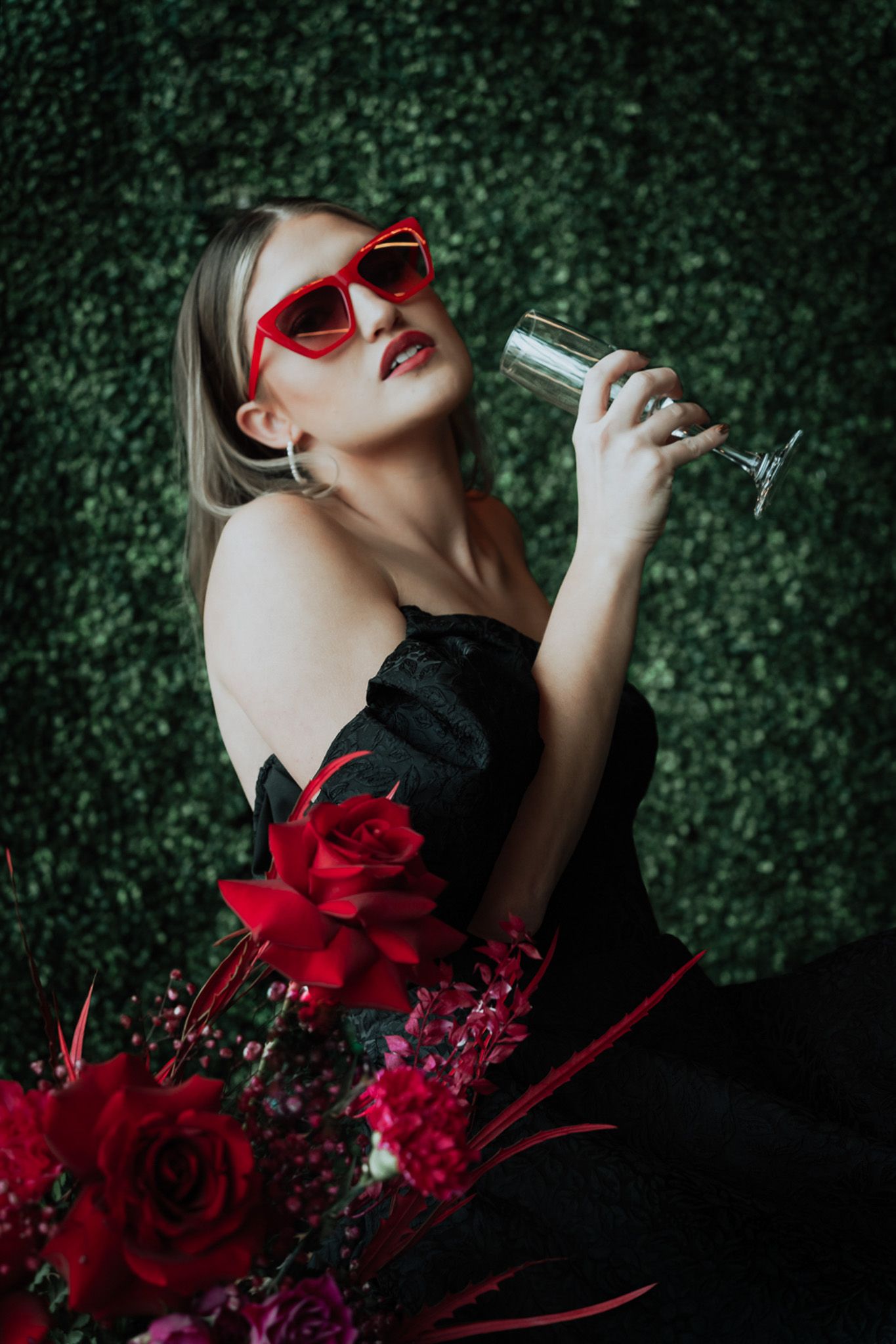 Glamorous portrait with red sunglasses and roses against green hedge backdrop.