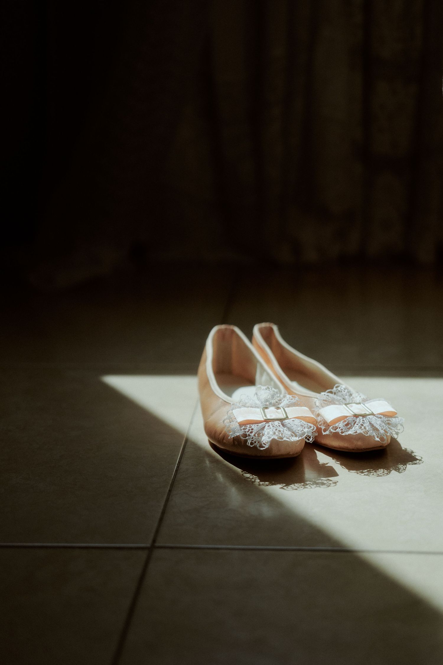 Elegant bridal flats with lace details cast dramatic shadows on a tiled floor.