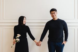 A couple in dark clothing poses playfully against a white wall with modern styling.