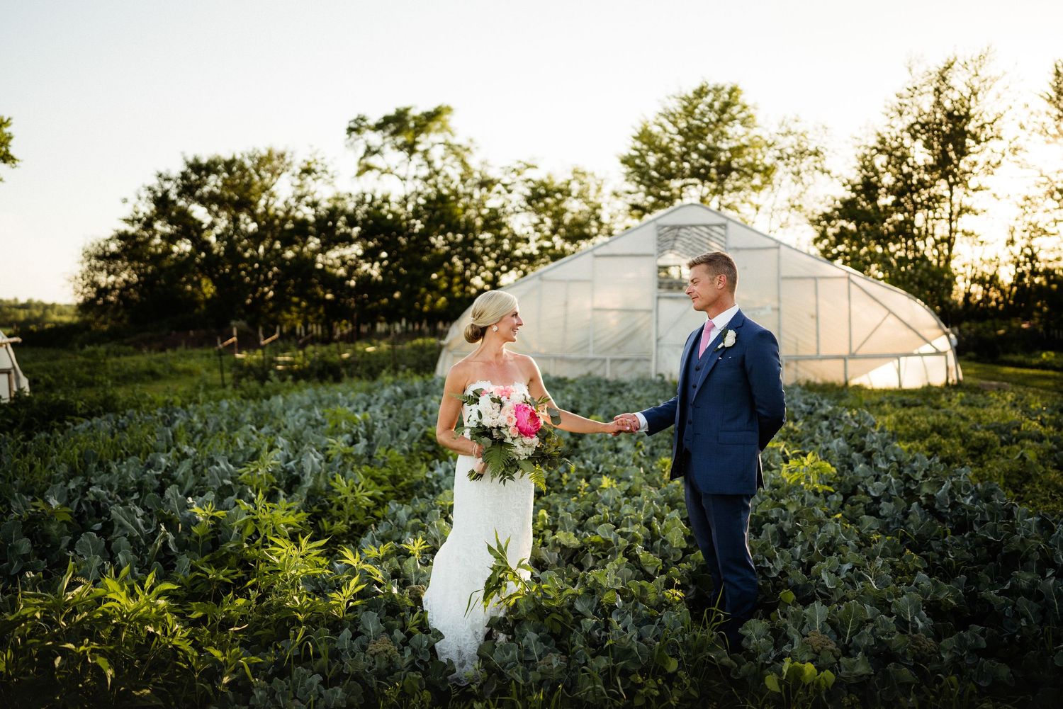 Wedding couple walking through lush garden field with greenhouse in background.