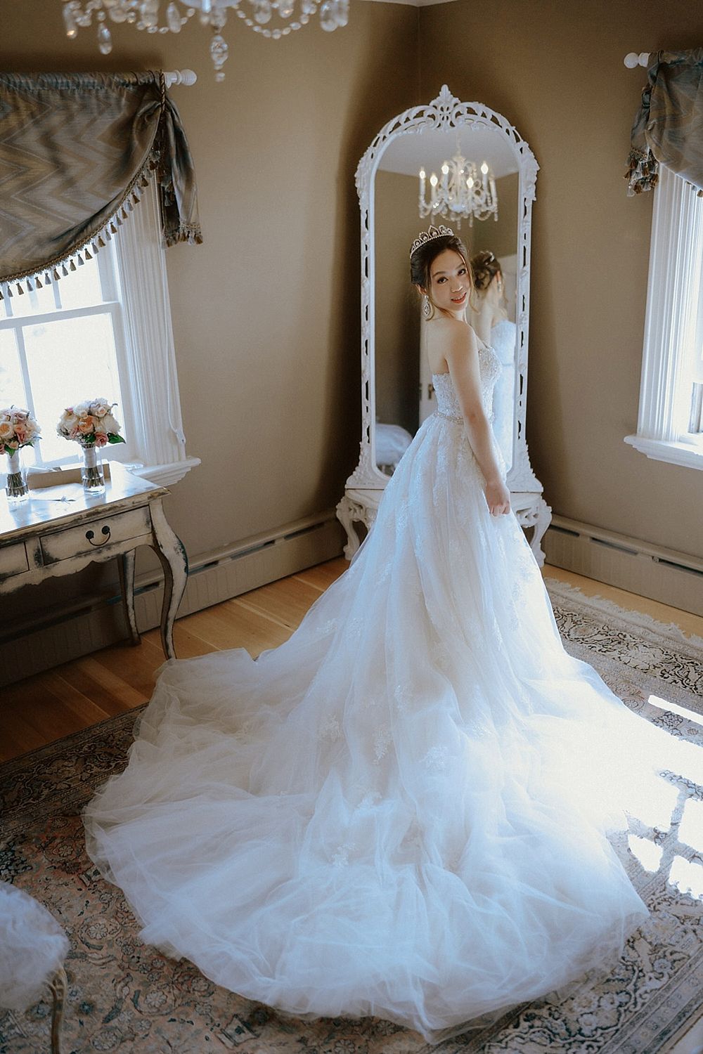 Wedding dress with flowing train displayed in front of an ornate white mirror in an elegant room.