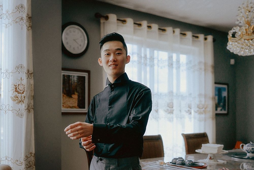 Elegant groom adjusting cuffs in formal attire by window.