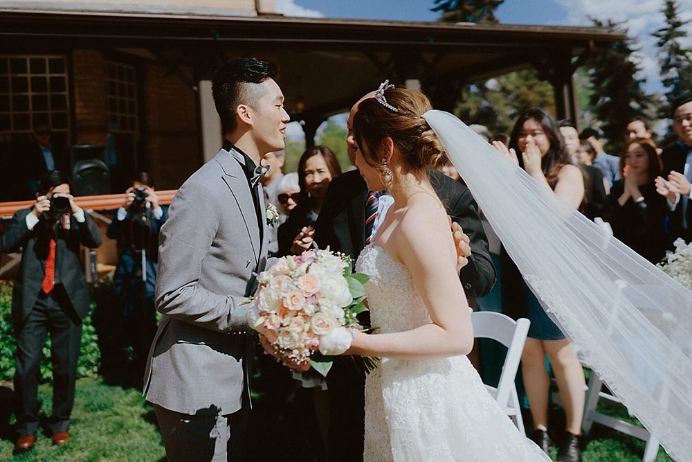 Outdoor wedding ceremony in progress with guests seated on white chairs near a rustic building.