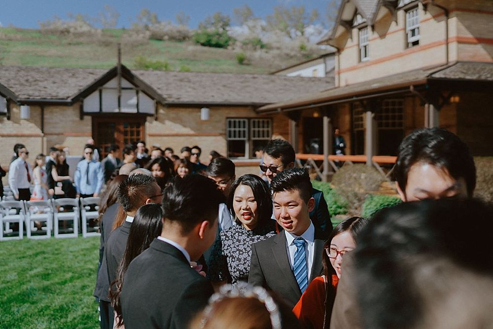 Wedding guests gather outside a rustic venue with Tudor-style architecture on a beautiful day.