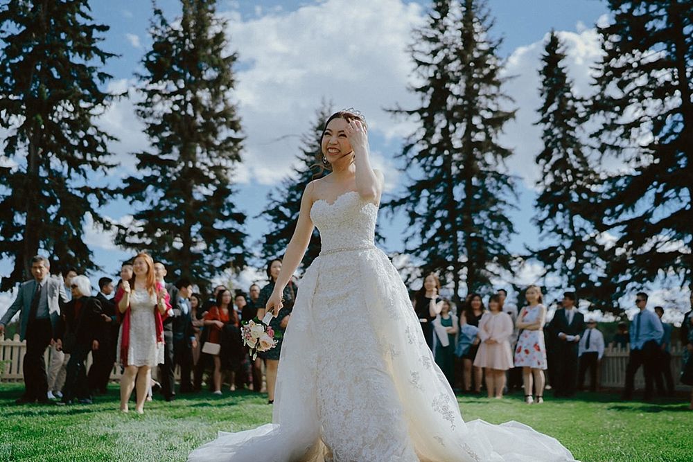 Wedding party celebrates on a green lawn surrounded by towering evergreen trees.