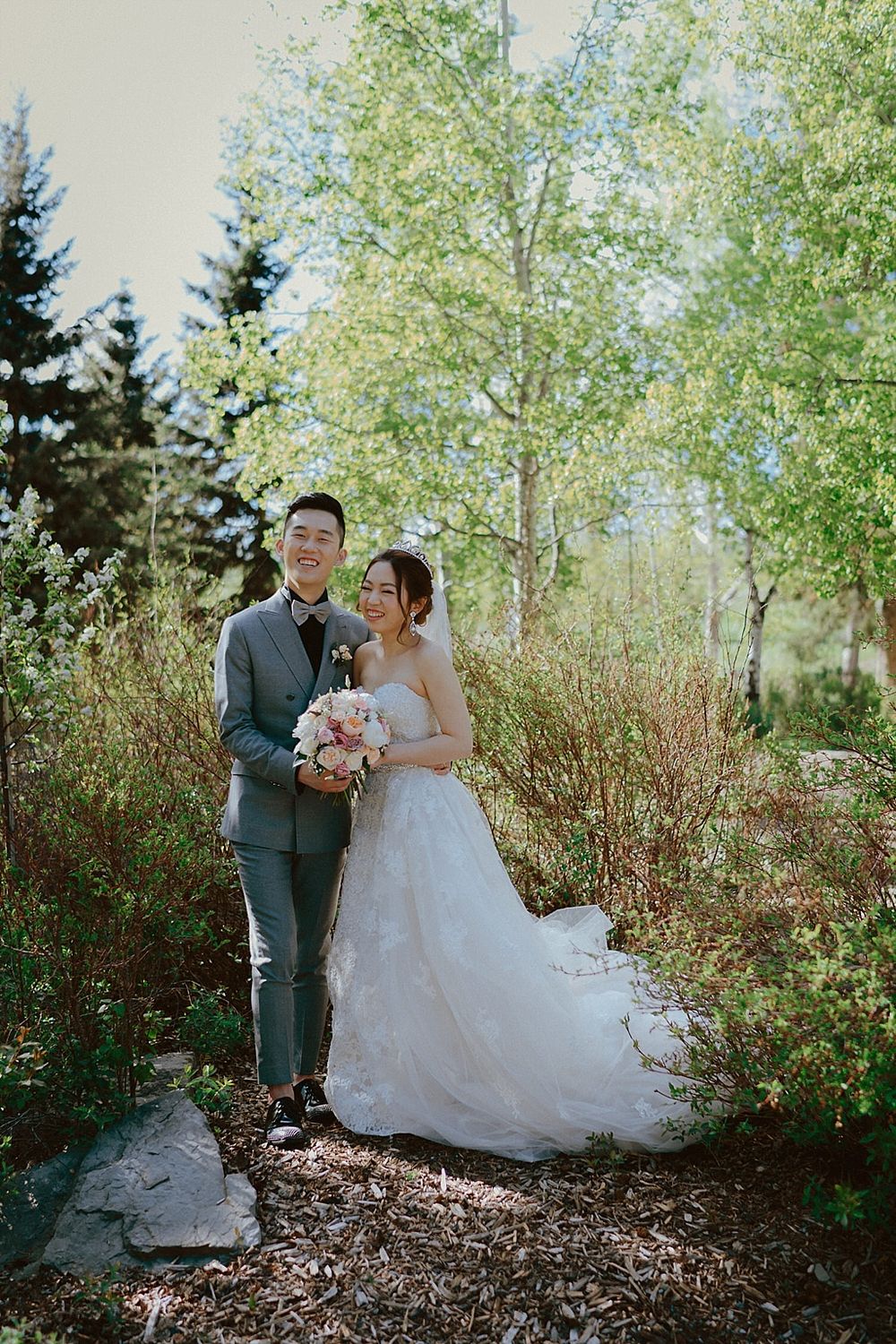 A wedding couple poses together in a garden setting with lush greenery and aspen trees.