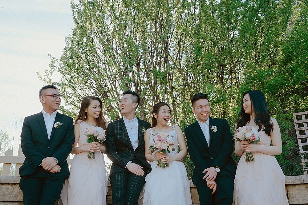 Wedding party in formal attire standing together outdoors with green trees in background.