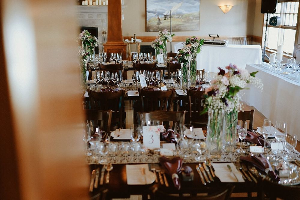 Elegant wedding reception table set with glass vases, candles and greenery in a rustic indoor venue.