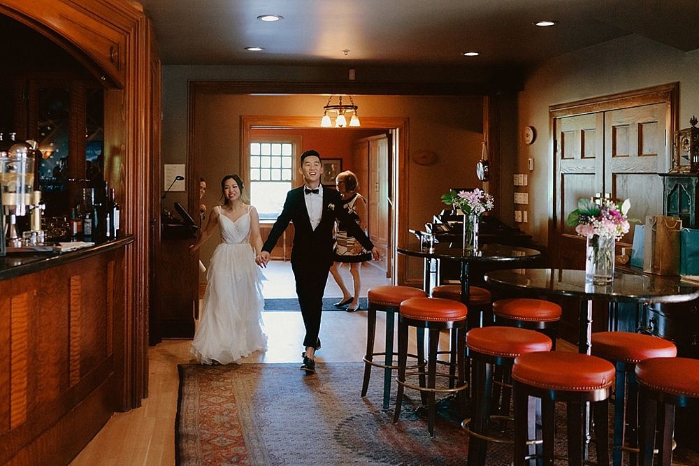 A wedding celebration in a rustic venue with a wooden bar and red barstools beside a sunlit doorway entrance.