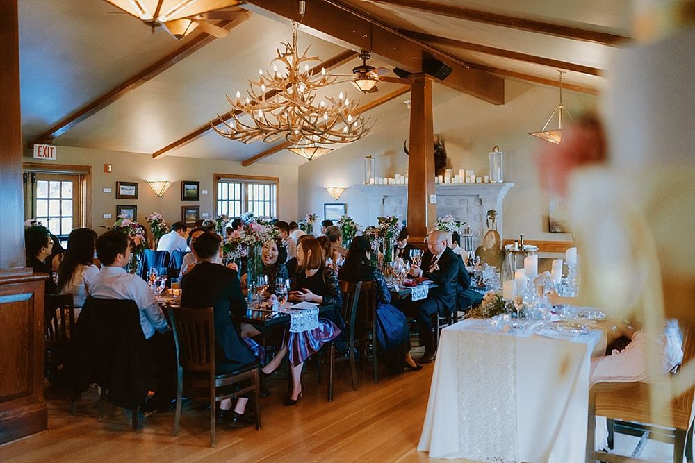 Wedding reception venue with rustic chandelier and guests seated at multiple tables.