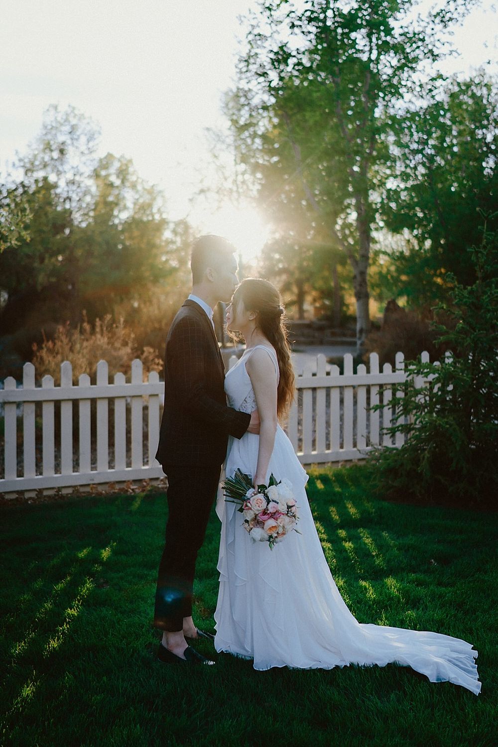 Couple in wedding attire embrace by a white picket fence during golden hour.