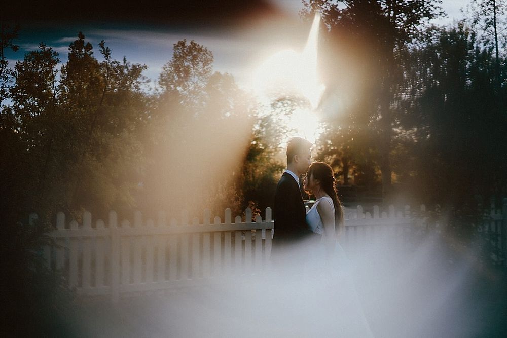 Dreamy backlit wedding portrait with white picket fence and trees in background.