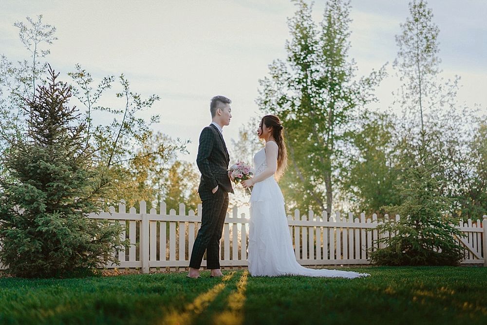 Wedding couple stands together against white picket fence with sunlight streaming through trees.