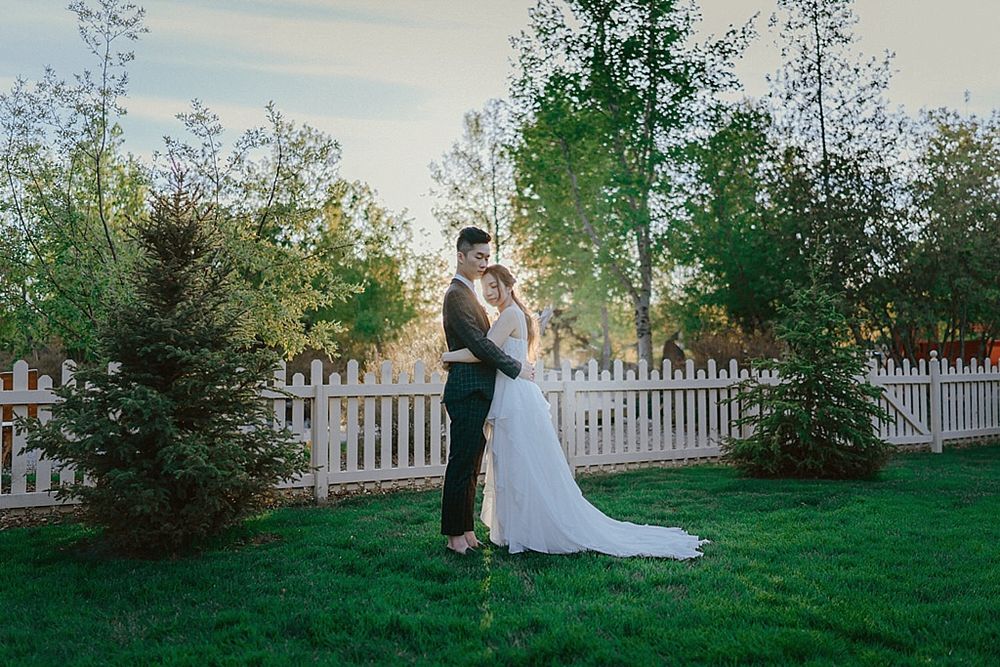 Wedding portrait of couple in elegant attire against white fence and manicured lawn.